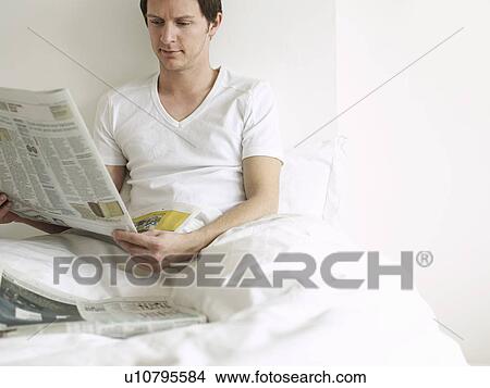 Picture - Young man reading newspaper in bed. Fotosearch