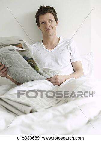 Stock Photo - Young man reading newspaper in bed looking to side and smiling. Fotosearch