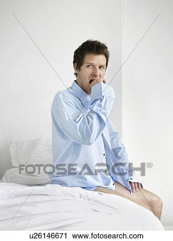 Stock Image - Young man sitting on bed and yawning. Fotosearch