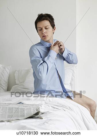 Stock Image - Young man sitting on bed buttoning up shirt and reading newspaper. Fotosearch