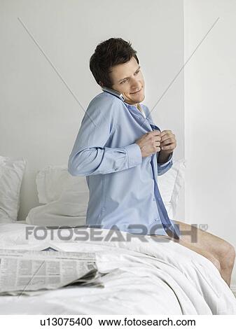 Young man sitting on bed buttoning up shirt and using phone View Large Photo Image Stock Image - Young man sitting on bed buttoning up shirt and using phone. Fotosearch