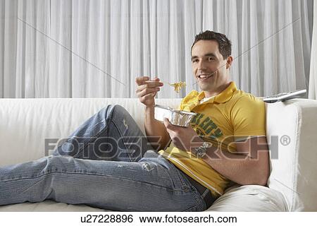 Stock Photograph - Young man sitting on sofa eating noodles from takeaway tray. Fotosearch