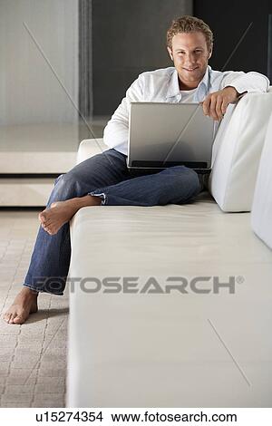 Young man sitting on sofa using laptop portrait View Large Photo Image Picture - Young man sitting on sofa using laptop portrait. Fotosearch