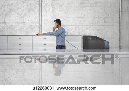 Stock Image - Young man standing by television set using phone side view. Fotosearch