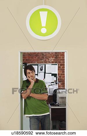 Stock Image - Young man standing in doorway of office with sign of exclamation mark above.. Fotosearch