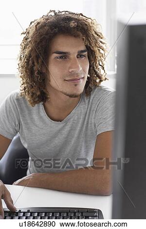 Stock Image - Young man using computer sitting at desk. Fotosearch