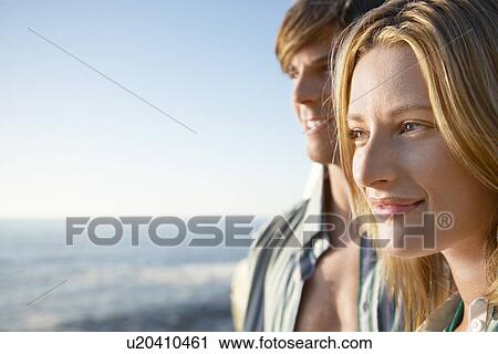 Stock Image - Young woman close up young man in background by ocean. Fotosearch