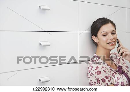 Stock Image - Young woman leaning on cabinet using mobile phone. Fotosearch