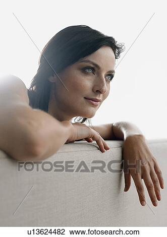 Stock Image - Young woman leaning on couch indoors. Fotosearch