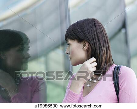 Stock Image - Young woman looking at reflection of self in building window. Fotosearch