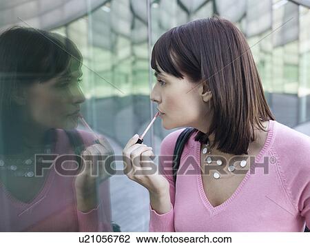 Stock Image - Young woman looking at reflection of self in building window applying lip gloss. Fotosearch