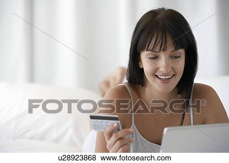 Stock Photo - Young woman lying on bed holding credit card whilst using laptop portrait. Fotosearch