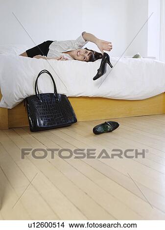 Picture - Young woman lying on bed throwing high heeled shoes on floor. Fotosearch