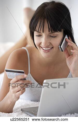 Young woman lying on bed with laptop holding credit card whilst using mobile phone portrait View Large Photo Image Stock Image - Young woman lying on bed with laptop holding credit card whilst using mobile phone portrait. Fotosearch