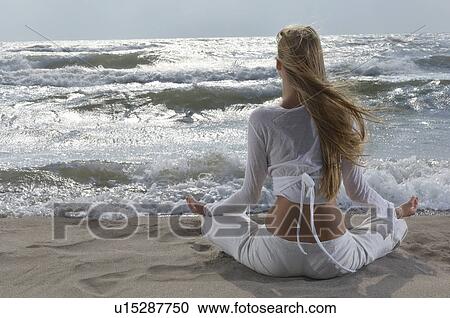 Stock Image - Young woman meditating on beach facing ocean back view. Fotosearch