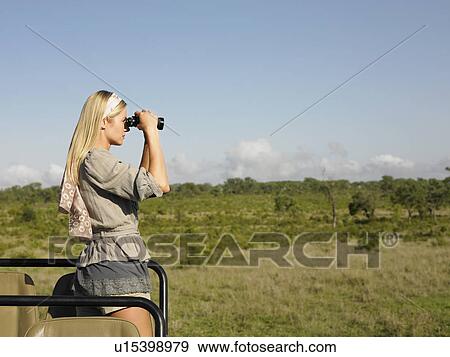 Stock Photo - Young woman on safari standing in jeep looking through binoculars side view. Fotosearch