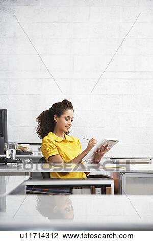 Stock Image - Young woman sitting at desk in office writing in notepad. Fotosearch