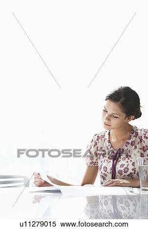Stock Photography - Young woman sitting at table holding and looking at piece of paper. Fotosearch