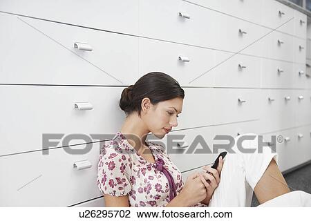 Stock Image - Young woman sitting by cabinet using mobile phone side view. Fotosearch