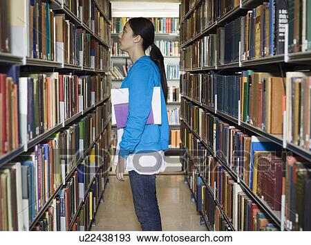 Stock Image - Young woman standing by bookshelf in library. Fotosearch
