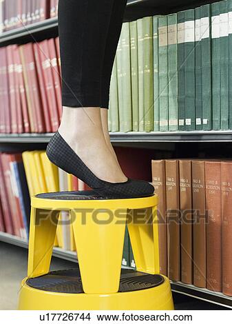 Picture - Young woman standing on stool reaching for book low section. Fotosearch