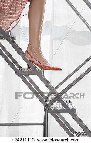 Stock Image - Young woman wearing high heeled shoes sitting on aluminium staircase low section side view. Fotosearch