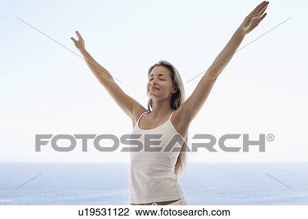Stock Image - Young woman with arms outstretched and eyes closed on beach. Fotosearch