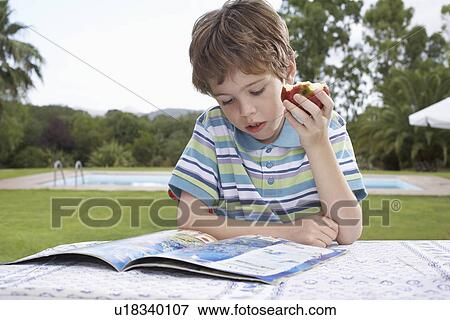 Boy (5-6) sitting at table reading and eating apple View Large Photo Image Stock Photo - Boy (5-6) sitting at table reading and eating apple. Fotosearch