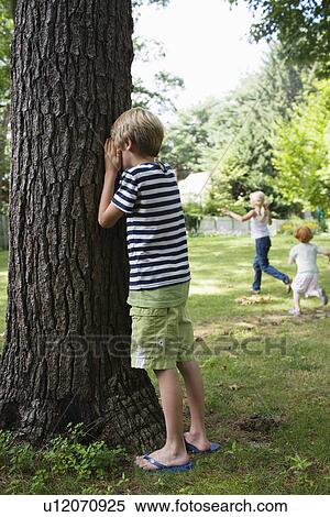Stock Photography - Boy (5-6) standing by tree with eyes covered children running in background. Fotosearch