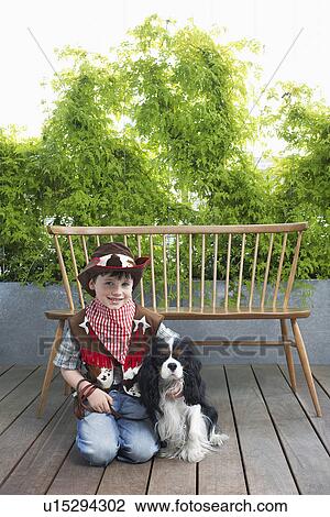 Stock Image - Boy (7-9) wearing cowboy costume kneeling with dog on decking. Fotosearch