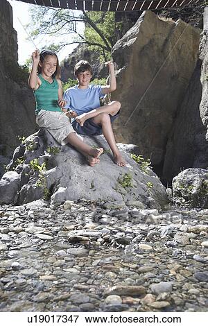 Stock Photo - Boy and girl (10-12) sitting on rock throwing stones. Fotosearch