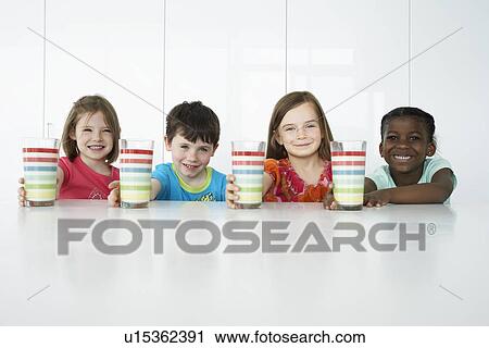Stock Image - Boys and three girls (5-6) sitting at table holding colourful glasses smiling portrait. Fotosearch