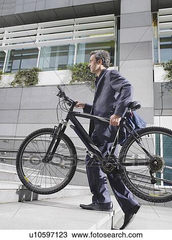 Stock Image - Business man carrying bicycle up steps side view. Fotosearch