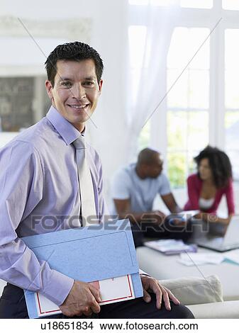 Business man holding document folder in living room couple sitting in background View Large Photo Image Picture - Business man holding document folder in living room couple sitting in background. Fotosearch