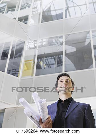 Business man holding rolled blueprints under arm in atrium of office building low angle view View Large Photo Image Picture - Business man holding rolled blueprints under arm in atrium of office building low angle view. Fotosearch