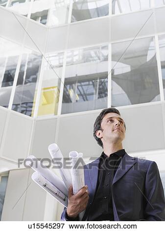 Stock Photo - Business man holding rolled blueprints under arm in atrium of office building low angle view. Fotosearch