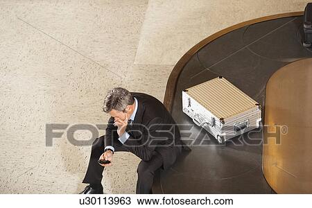Business man looking at mobile phone sitting on luggage carousel in airport View Large Photo Image Stock Image - Business man looking at mobile phone sitting on luggage carousel in airport. Fotosearch