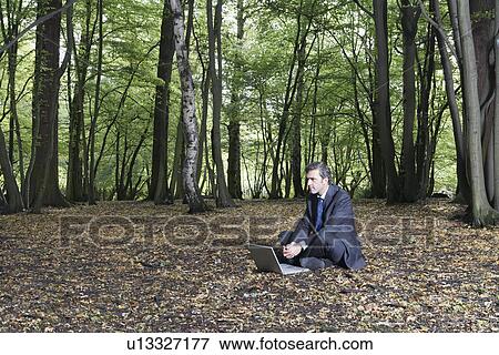 Stock Photo - Business man sitting on ground using laptop in middle of forest. Fotosearch