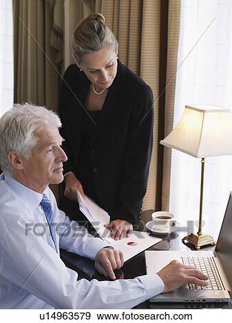 Stock Photo - Business man using laptop business woman sitting on desk looking at documents elevated view. Fotosearch