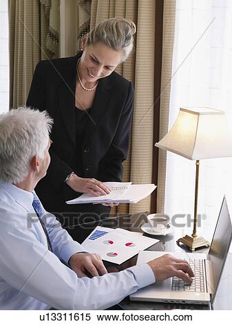 Stock Photography - Business man using laptop business woman sitting on desk holding documents elevated view. Fotosearch