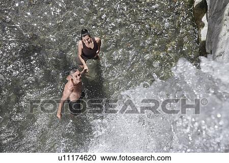 Stock Image - Couple in river under waterfall looking up elevated view. Fotosearch