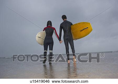 Couple of surfers carrying surfboards walking on beach back view View Large Photo Image Stock Photograph - Couple of surfers carrying surfboards walking on beach back view. Fotosearch