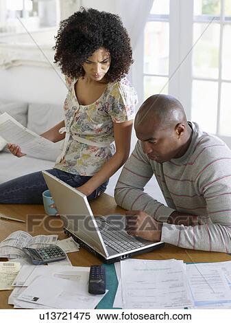 Stock Photography - Couple sitting at table using laptop and holding bills elevated view. Fotosearch