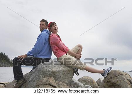 Stock Photography - Couple sitting back to back on rocks by ocean portrait. Fotosearch