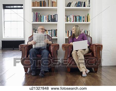 Couple sitting in armchairs man reading newspaper woman using laptop View Large Photo Image Stock Photo - Couple sitting in armchairs man reading newspaper woman using laptop. Fotosearch