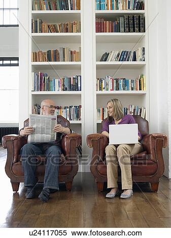 Stock Photography - Couple sitting in armchairs man reading newspaper woman using laptop. Fotosearch