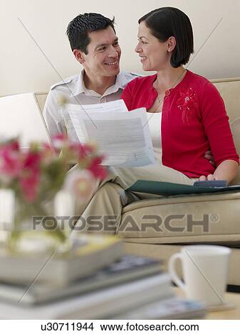 Picture - Couple using laptop and holding documents sitting on sofa. Fotosearch
