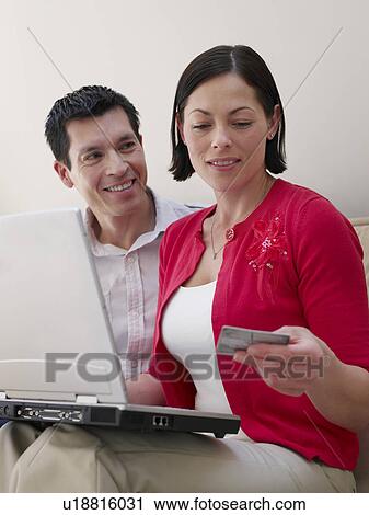 Stock Image - Couple using laptop sitting on sofa woman holding credit card. Fotosearch