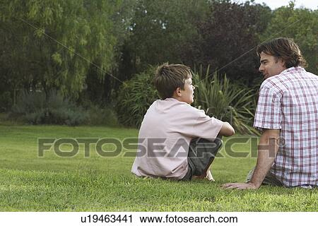 Stock Image - Father and son (10-12) sitting on lawn and talking. Fotosearch