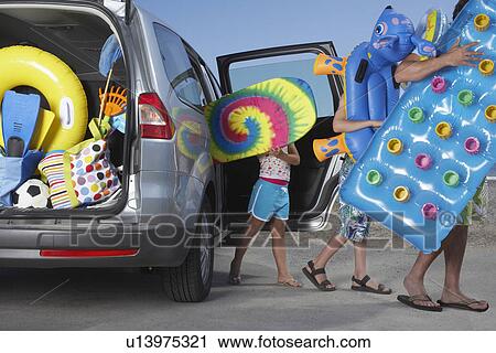 Stock Image - Father and two children (6-11) unloading beach accessories from car. Fotosearch